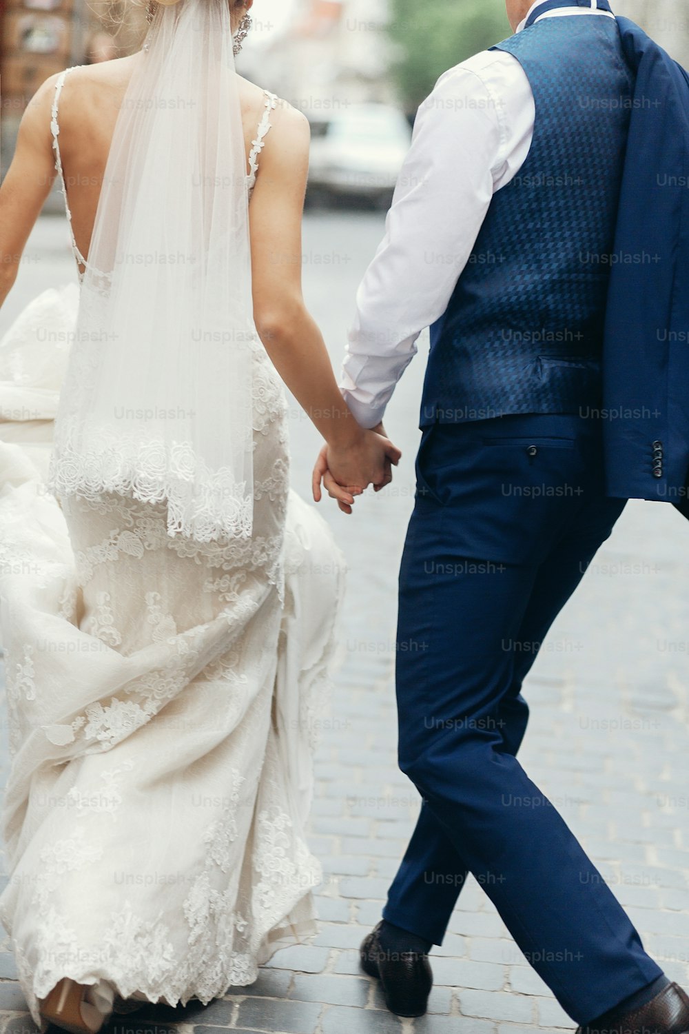 Elegante pareja de recién casados tomados de la mano mientras caminan por la ciudad después de la ceremonia de la boda, hermosa novia rubia caminando con el novio guapo