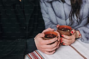 Stylish couple holding cups with hot tea in hands on wooden porch in winter snowy mountains. Happy romantic family with drinks. Holiday getaway together. Romantic moments