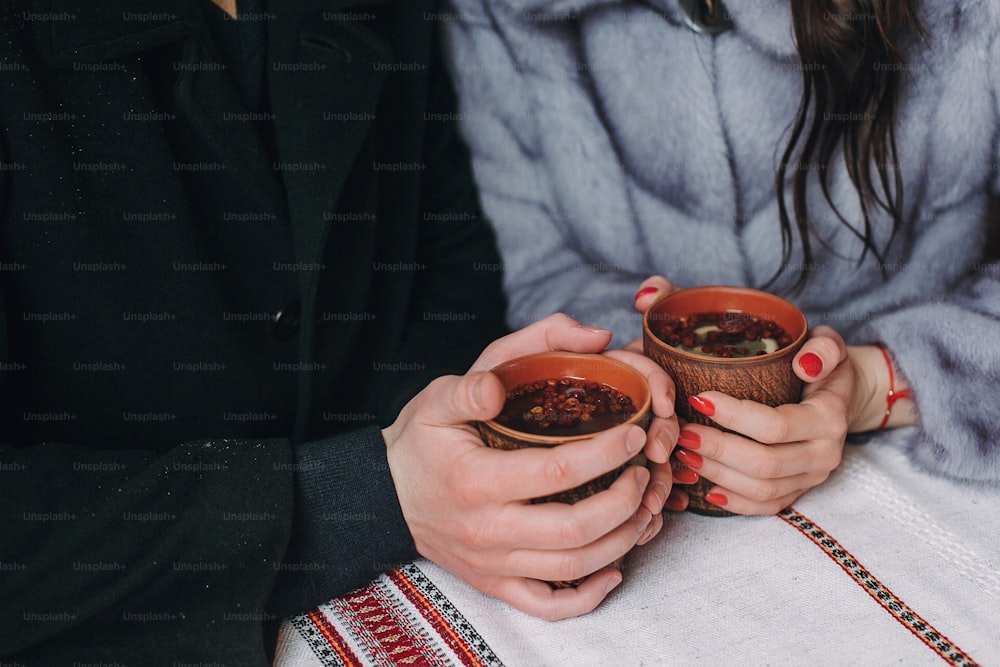 Stylish couple holding cups with hot tea in hands on wooden porch in winter snowy mountains. Happy romantic family with drinks. Holiday getaway together. Romantic moments