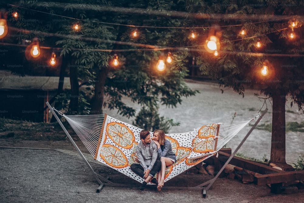 stylish hipster couple cuddling and relaxing in hammock under retro lights in evening summer park. man hugging woman and resting in forest. space for text. rustic love concept