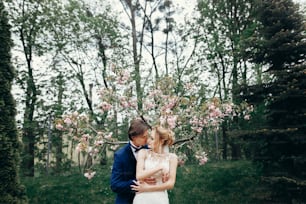 Stilvolle Braut und Bräutigam umarmen und küssen sich im Park zwischen Magnolienblüten. Leidenschaftliches Luxus-Hochzeitspaar umarmt sich. romantischer sinnlicher Moment.
