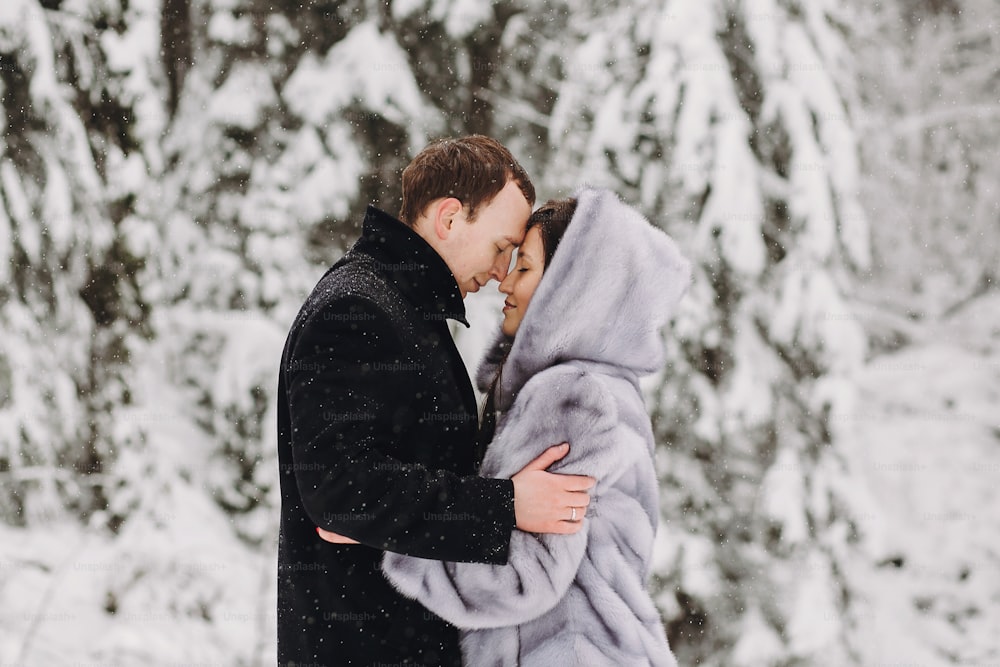 Stilvolles Paar, das sich im Winter in schneebedeckten Bergen umarmt. Glücklicher romantischer Mann und Frau in Luxuskleidung, die sich sanft am Wasserfall im Schnee umarmen. Gemeinsamer Urlaub