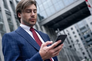 Confident businessman. Confident young man in full suit is talking by his smart phone and looking away while standing outdoors with cityscape in the background