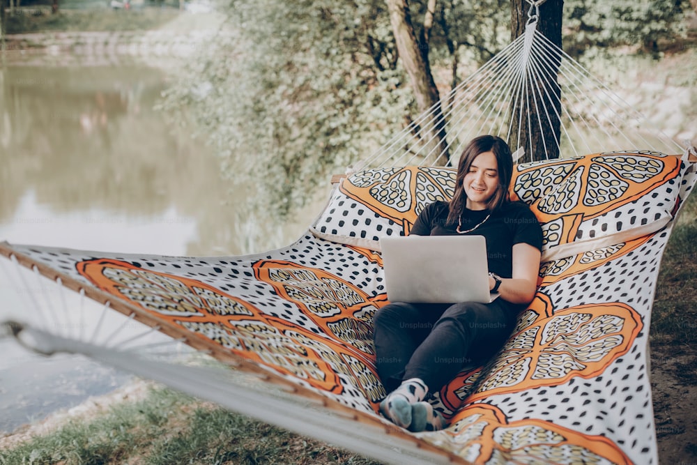 girl working on laptop while lying on hammock in sunny summer park. freelancer woman hipster with laptop relaxing and smiling. freelance concept. student study. summer vacation