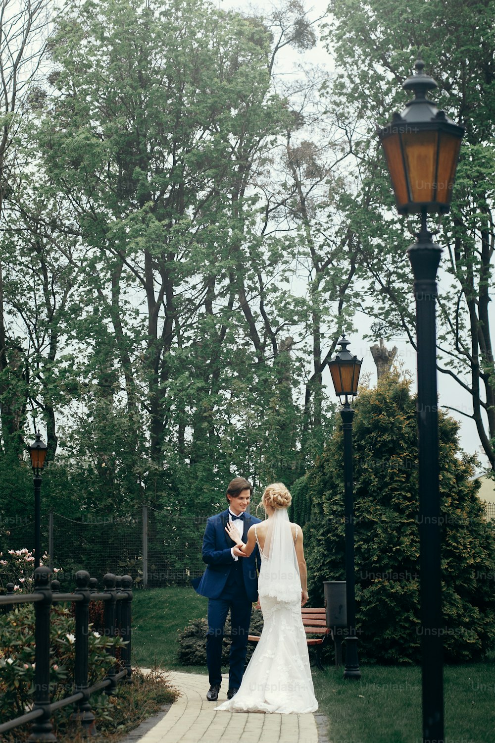 Stilvolles Brautpaar tanzt im Park.  Glückliches Luxushochzeitspaar, das zwischen Bäumen spazieren geht und lächelt. Mann im blauen Anzug und Frau im weißen Kleid an der Rezeption im Freien