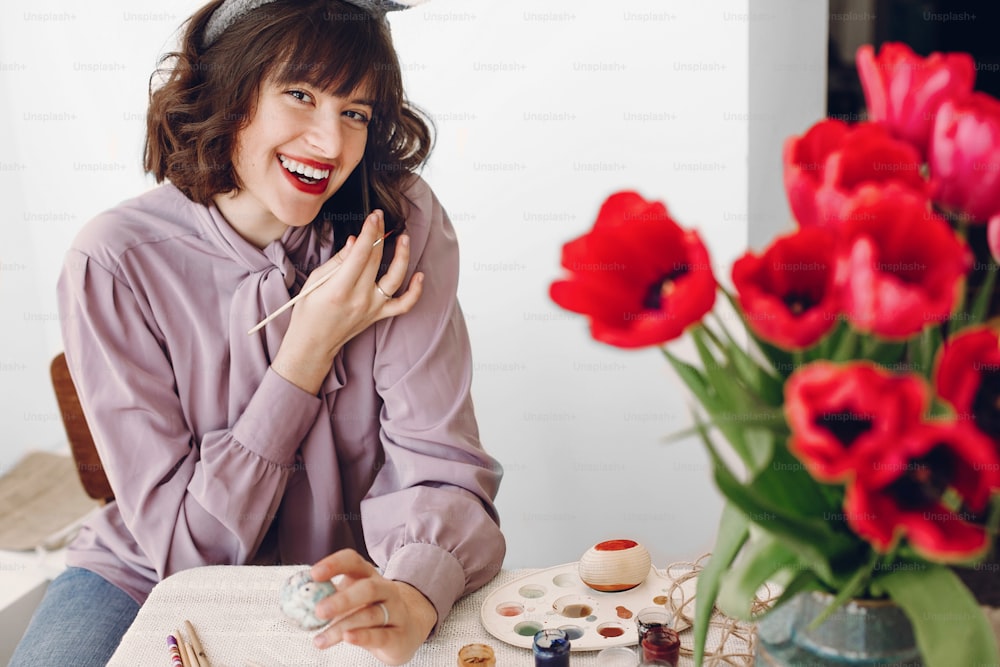 happy easter greetings. beautiful stylish girl in bunny ears smiling and painting easter eggs on rustic table with colorful paint and tuilips in home with space for text. woman talking on phone