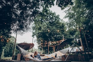 세련된 힙스터 커플은 저녁 여름 공원의 복고풍 조명 아래 해먹에서 껴안고 휴식을 취하고 있습니다. 여자를 껴안고 숲에서 쉬고 있는 남자. 텍스트 공간. 소박한 사랑 개념
