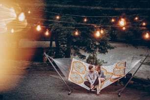 Coppia hipster romantica si gode il riposo su un'amaca al resort del parco, donna carina sdraiata con l'uomo bello, luci sullo sfondo