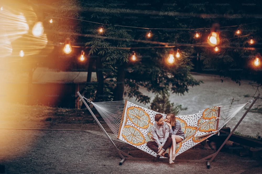ロマンチックなヒップスターのカップルは、公園のリゾート地でハンモックの上で休息を楽しみ、ハンサムな男と横たわっているかわいい女性、背景にライトがあります