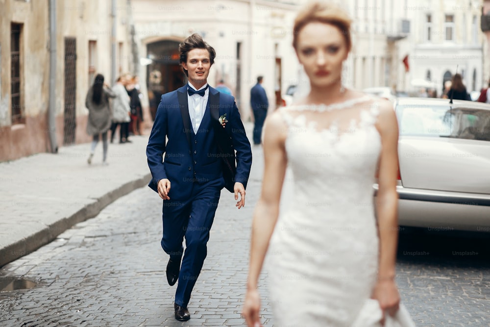 mariée élégante marchant et marié courant après elle dans la rue de la ville. heureux couple de mariage de luxe s’amusant et dansant. moment romantique.