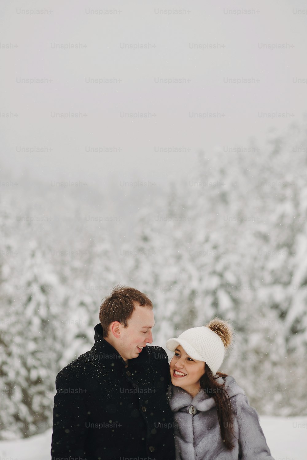Stylish couple in love hugging in snowy mountains. Portraits of happy family gently embracing and smiling in winter mountains and forest. Holiday getaway together