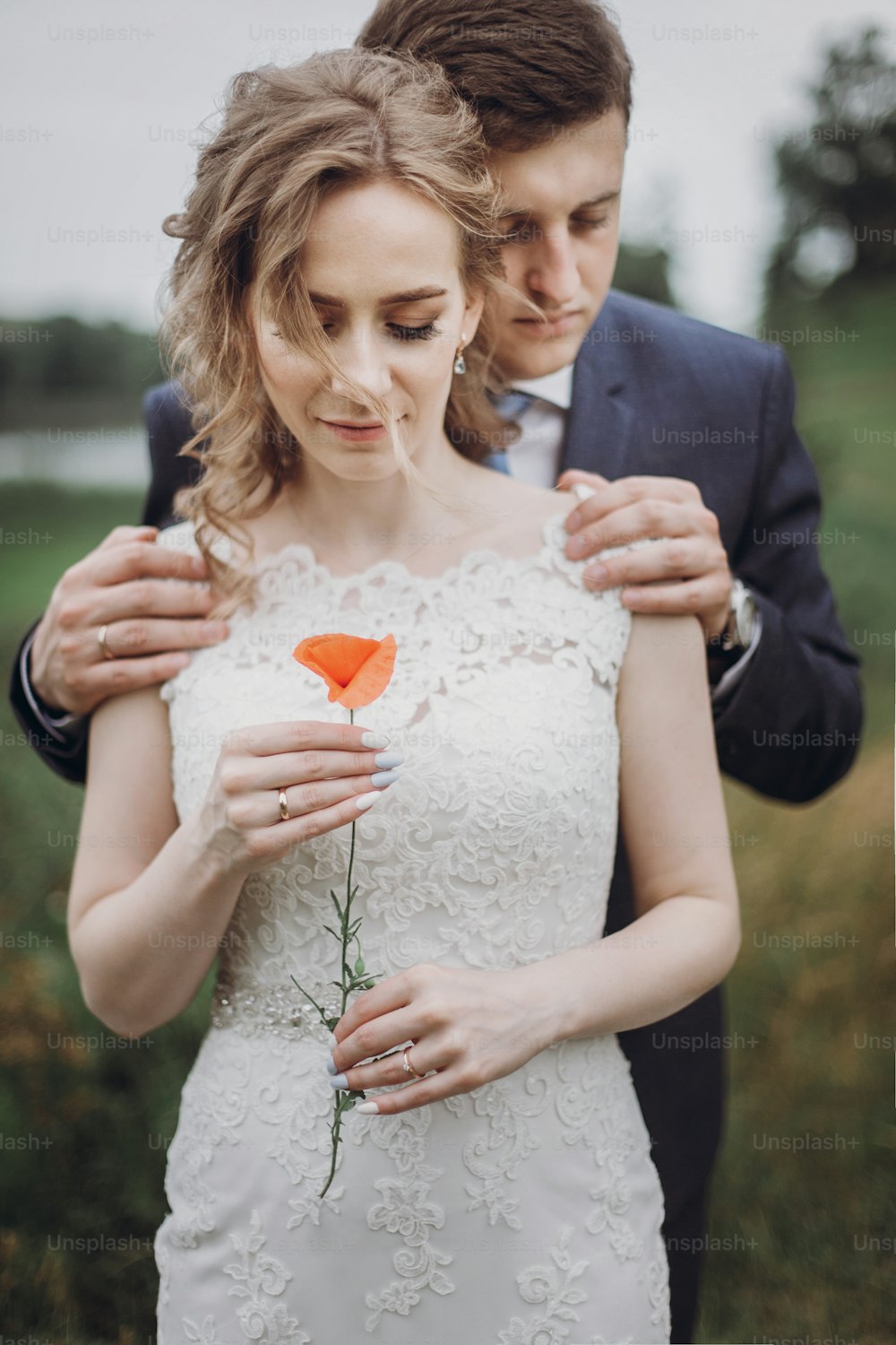 Gorgeous blonde bride in elegant lace wedding dress holding a flower while being hugged from behind by handsome groom, newlywed couple portrait