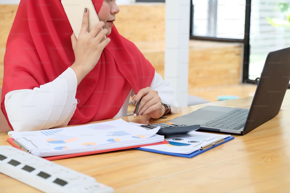 テーブルで働き、スマートフォン通話を使用するイスラムの女性。