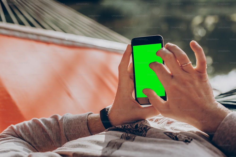 Hombre hipster sosteniendo un teléfono inteligente con pantalla vacía con espacio para texto, y relajándose en una hamaca en un parque de verano soleado. Maqueta. chico mirando el teléfono en blanco, navegando por internet en línea