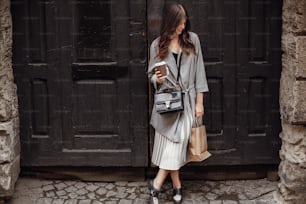 Gorgeous young woman with cup of coffee and bag, smiling and standing at old wooden door in city street. Stylish happy hipster girl with beautiful hair and perfect smile shopping