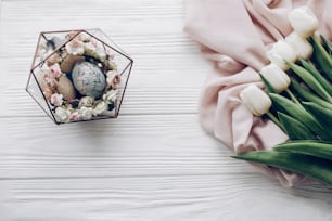 happy easter greeting card. stylish easter eggs with floral ornaments in nest of flowers and tulips on rustic white wooden background flat lay. space for text, top view