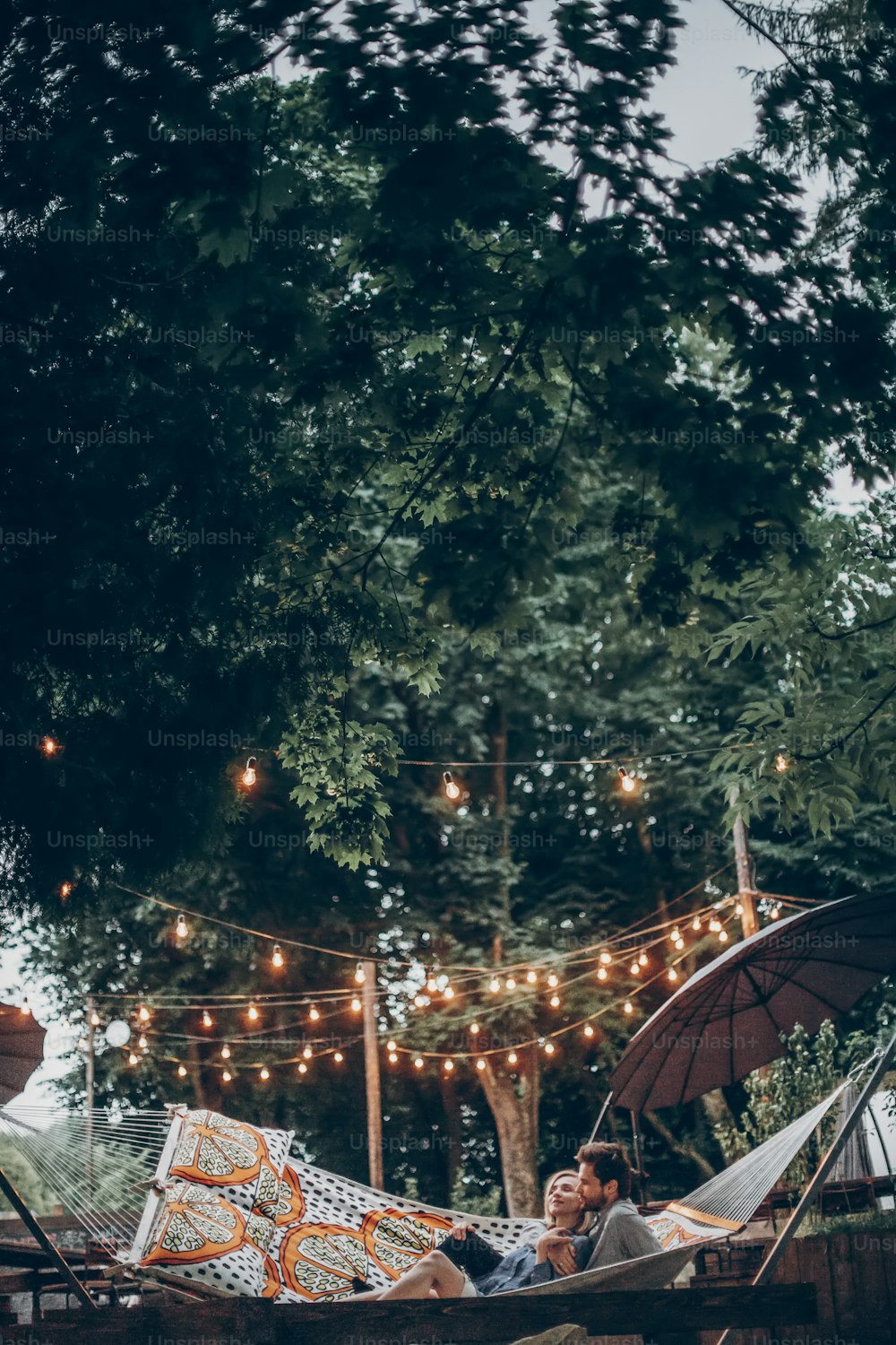 stylish hipster family couple cuddling and relaxing in hammock under retro lights in evening summer park. rustic man and woman embracing and resting in forest. space for text. atmospheric moment