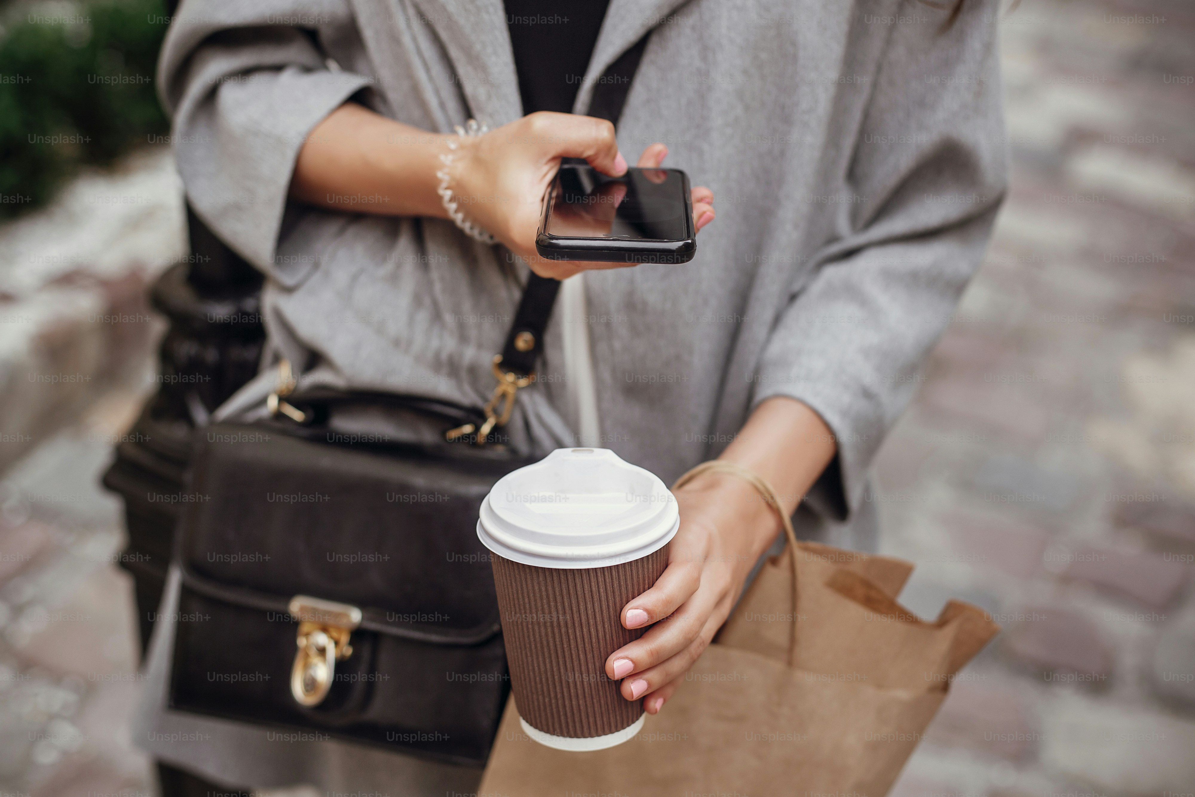 Stylish hipster girl holding  phone, and taking photo of coffee cup, bag, and feet in city street. Gorgeous happy young woman enjoying time and posting on social media