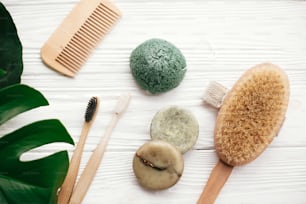 Zero waste flat lay. Natural solid shampoo bar, bamboo toothbrushes, wooden brush,  deodorant cream and konjaku sponge on white wood with green monstera leaves. Eco products plastic free