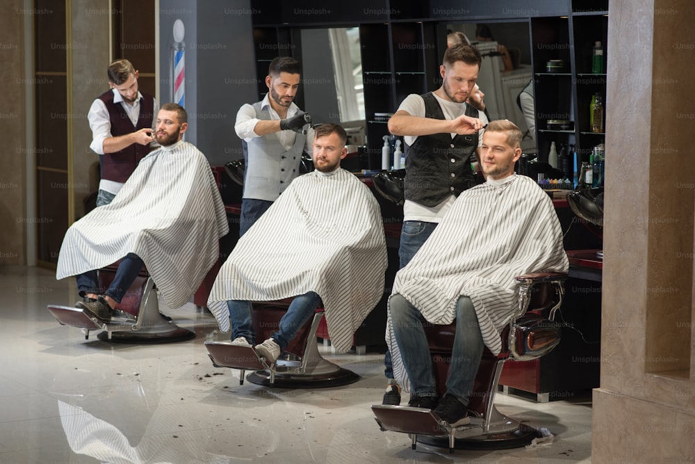Prozess des Stylings und der Pflege von Männerhaarschnitten im Friseursalon. Drei professionelle und selbstbewusste Friseure, die stehen und den Männern die Haare schneiden. Männliche Kunden, die auf Stühlen sitzen und Haarschnittkittel tragen.