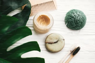 Eco friendly natural bamboo toothbrushes, shampoo bar, toothpaste in glass, wooden brush and konjaku sponge on white wood with green monstera leaves. Zero waste concept, plastic free