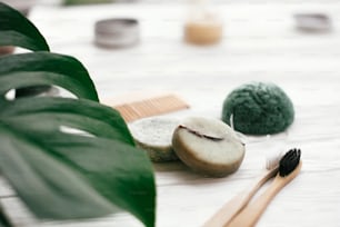Zero waste solid shampoo bar, bamboo toothbrushes, wooden brush and konjaku sponge on white wood with green monstera leaves. Eco friendly natural products plastic free