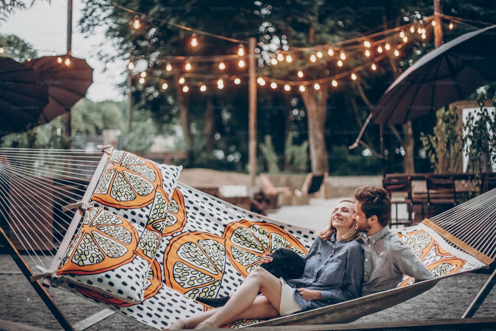Romantic hipster couple enjoy rest on a hammock at park resort, cute woman lying with handsome man, lights in the background