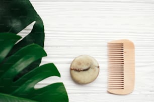 Natural eco friendly solid shampoo bar, wooden brush,  green  conditioner, soap on white wood with green monstera leaves.  Eco products plastic free. Zero waste. Hair care
