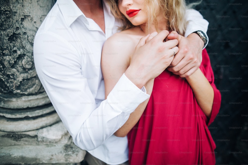 beautiful happy couple in love posing at old building in sunny street. stylish hipster groom and blonde bride in red dress gently hugging, professional dancers. romantic moments in summer