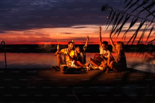 Group of young friends enjoying at the lake at night. They sitting around the fire singing and having fun at camping.