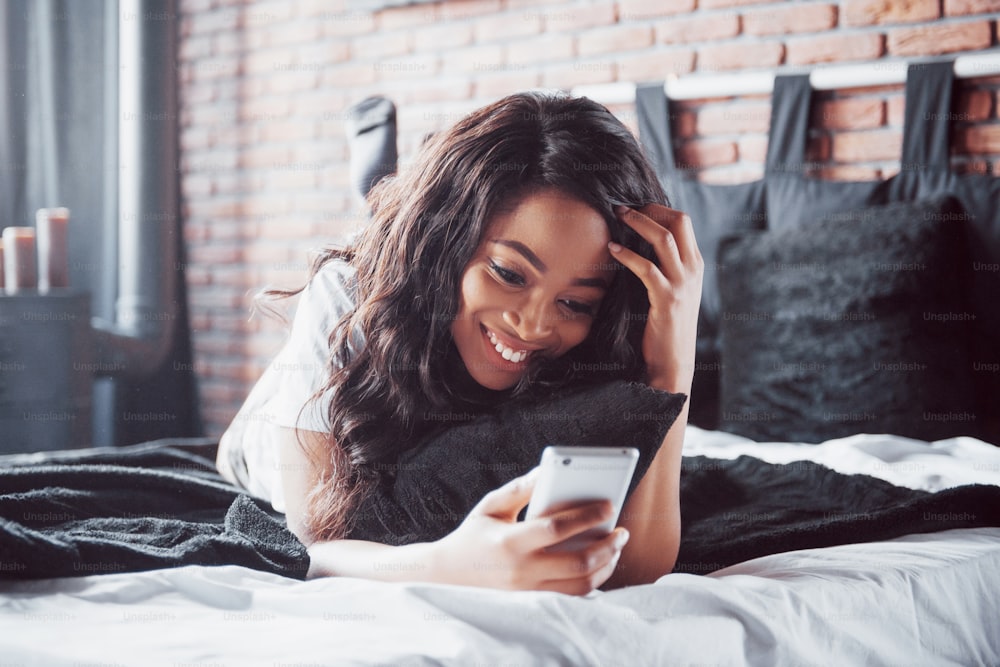 Retrato de una mujer hermosa que se despierta en su cama y mira el teléfono. Revisa las redes sociales, envía sms. La chica lleva una camiseta.