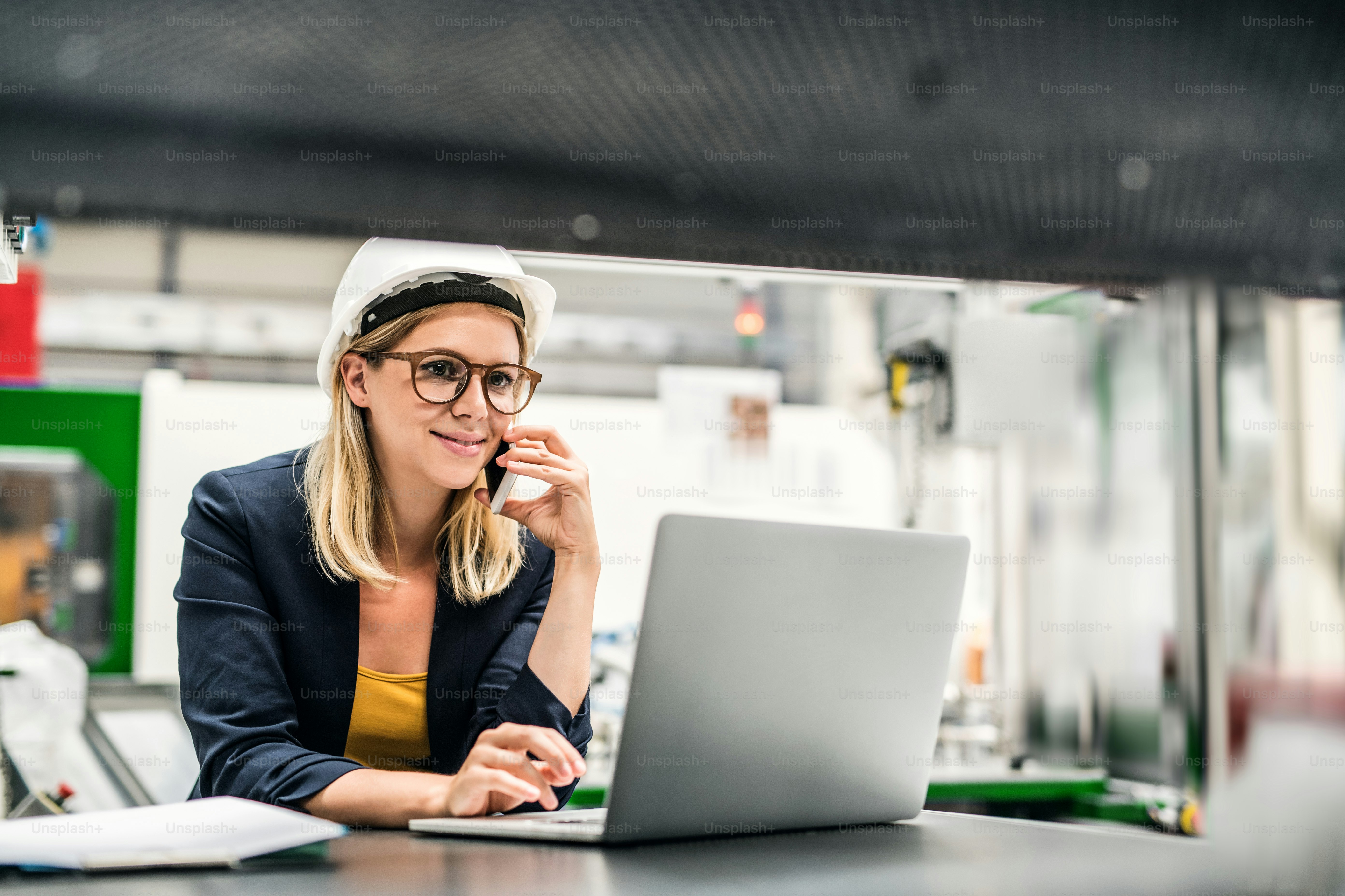 A portrait of a young industrial woman engineer in a factory, using laptop and smartphone.