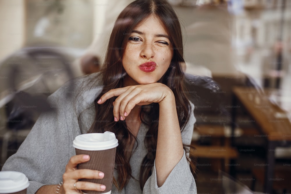Gorgeous young woman showing kiss, sitting in cafe with cup of coffee and looking through window on city street. Stylish hipster girl with beautiful hair kissing and smiling