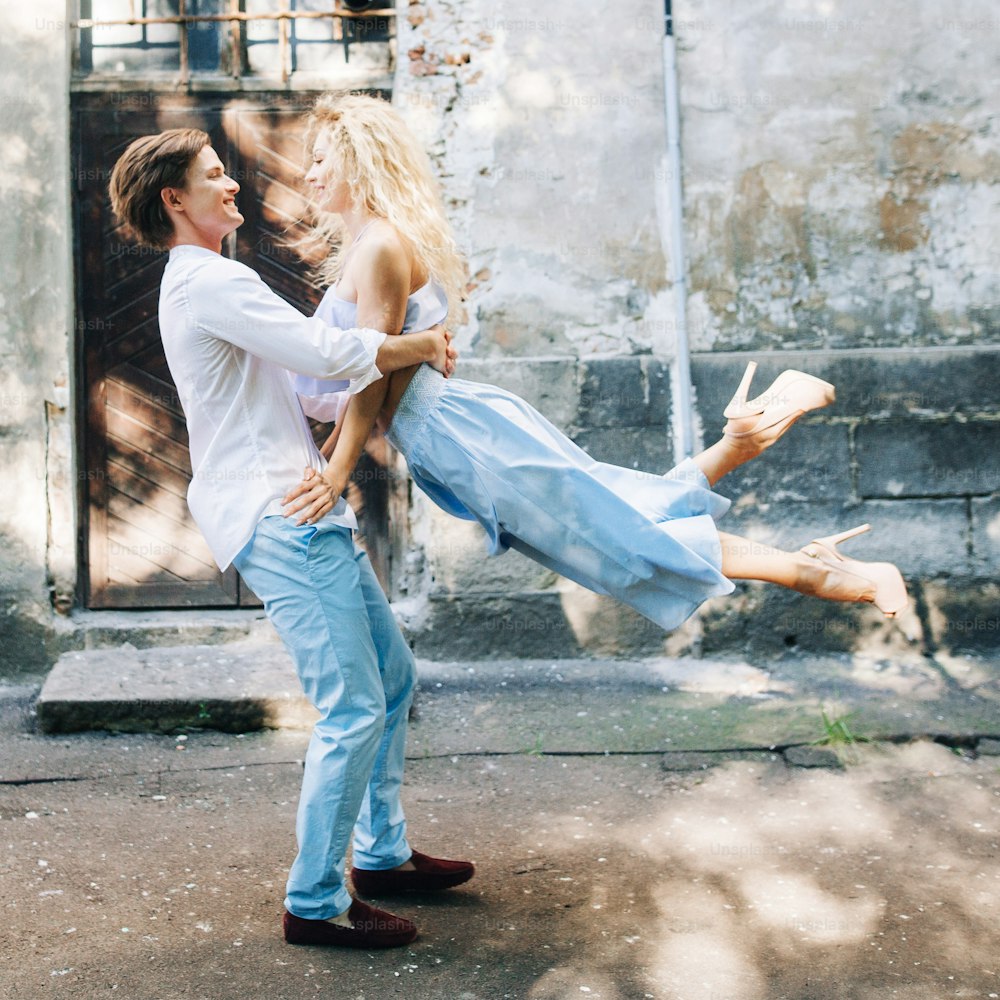 Schönes, glückliches verliebtes Paar beim Wandern und Tanzen in der sonnigen grünen Straße. Stilvoller Hipster-Bräutigam und blonde Braut haben Spaß. Romantische Momente im Sommer City Street im Urlaub