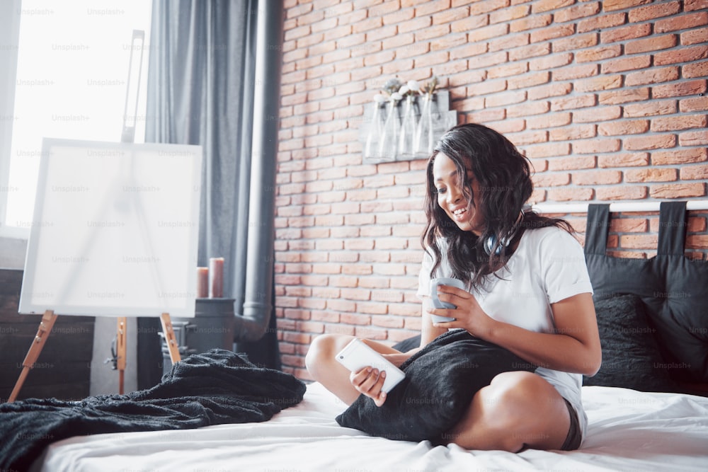 Retrato de mulher bonita acordando em sua cama e olha para o telefone. Verifique as redes sociais, envie sms. A menina está vestindo uma camiseta.