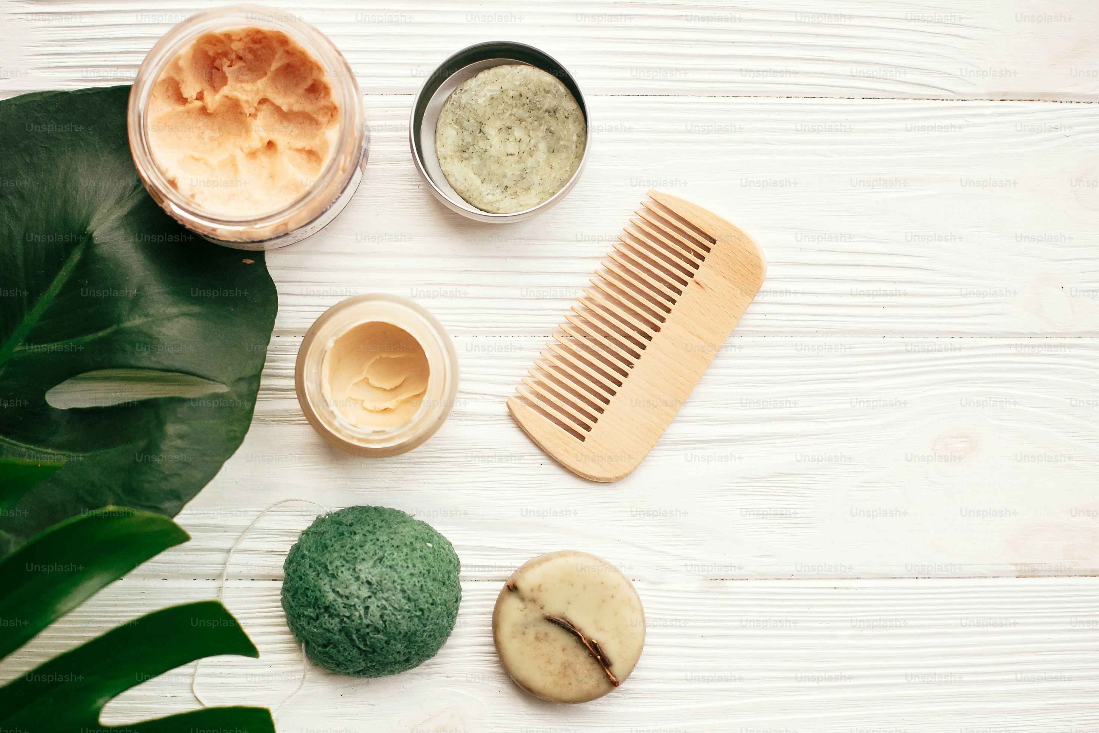 Skincare products, a wooden comb, and a green leaf on a white wooden surface