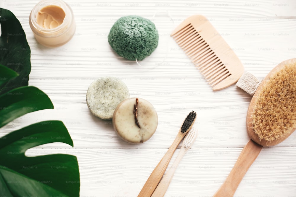 Zero waste flat lay. Natural solid shampoo bar, bamboo toothbrushes, wooden brush,  deodorant cream and konjaku sponge on white wood with green monstera leaves. Eco products plastic free
