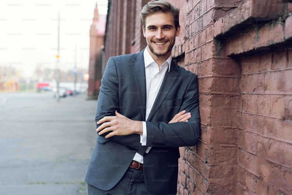 Gerente masculino joven sonriente vestido formalmente apoyado en una pared al aire libre