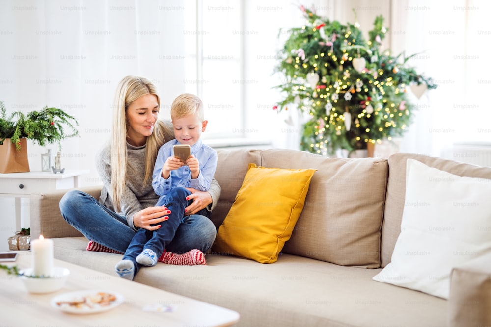 Una madre e un bambino piccolo con smartphone seduti su un divano a casa nel periodo natalizio, giocando.