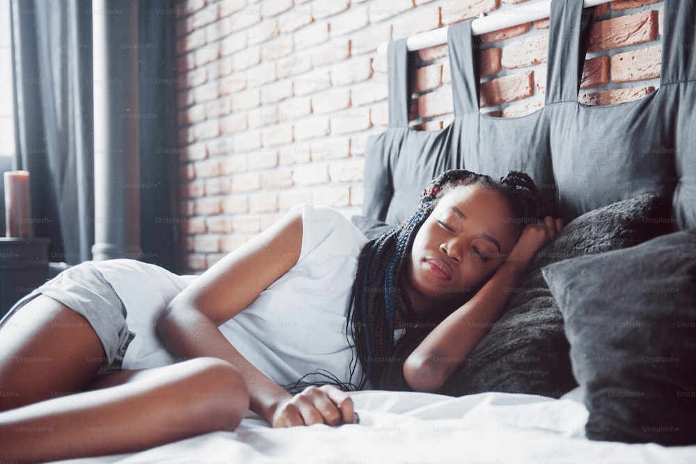 아름다운 아프리카 여자가 하얀 침실에서 침대에 누워 잔다. 좋은 아침, 집에서 휴가. 소녀는 머리에 땋은 머리를 한 티셔츠를 입고  있습니다. 사진 – Unsplash의 베개 이미지