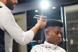 Side view of serious man sitting on chair and getting trendy haircut in barber shop. Barber in white shirt keeping clipper and correcting form of hair. Concept of hairstyling and trimming.