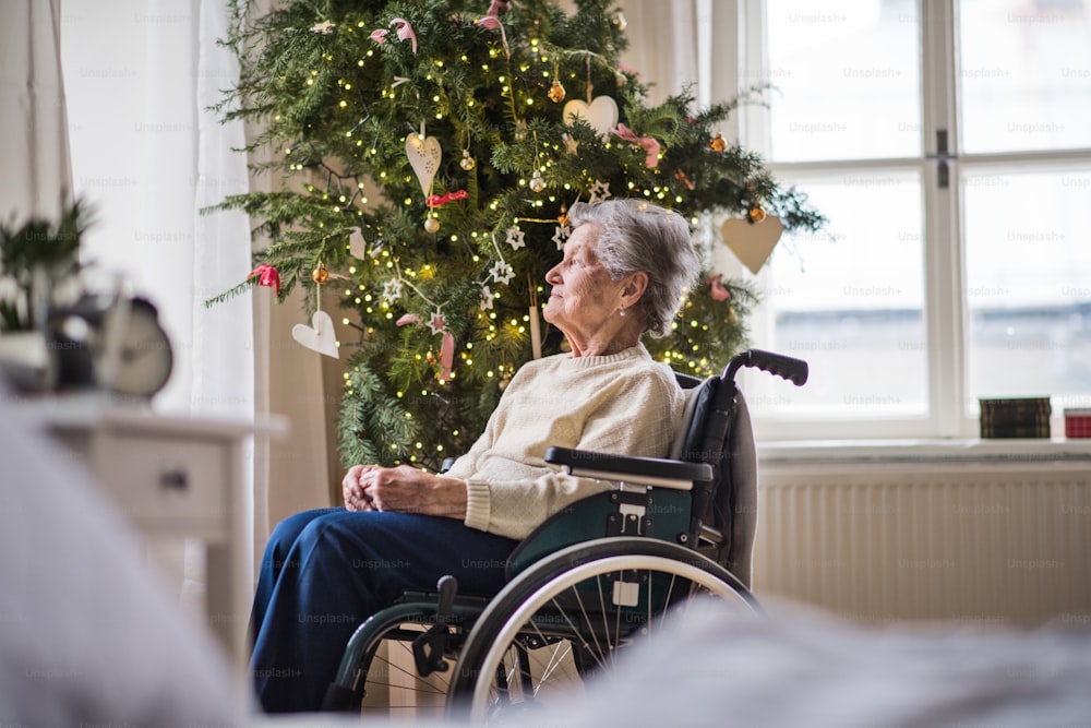Una donna anziana sola in sedia a rotelle a casa nel periodo natalizio, guardando fuori da una finestra.