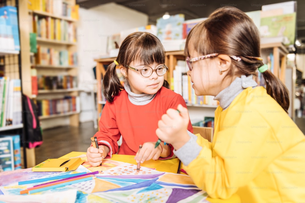 En la biblioteca infantil. Lindas hermanas atractivas con síndrome de Down coloreando imágenes en la biblioteca de los niños juntas