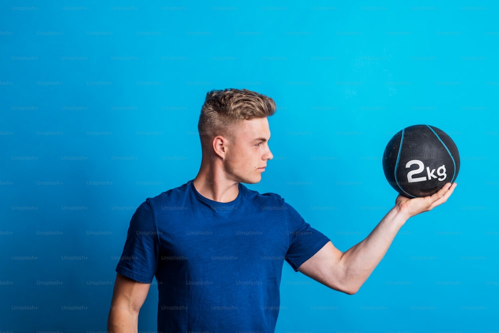 Portrait of a young man holding heavy medicine ball in one hand in a studio, standing against a blue background.