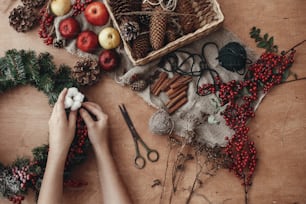 素朴なクリスマスリースフラットレイ。素朴な木製の背景にモミの枝、赤い果実、松ぼっくり、糸、はさみ、シナモンに綿を置く手。休日のワークショップでリースを作る