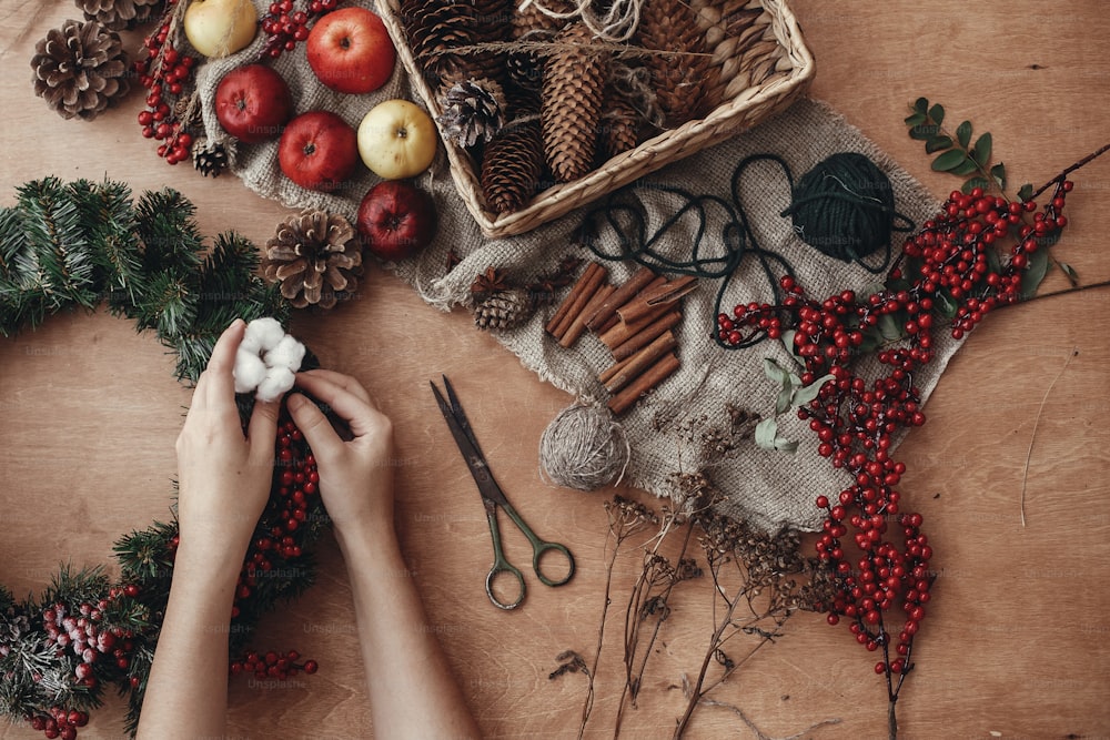 Rustic Christmas wreath flat lay. Hands putting cotton on fir branches, red berries and pine cones,thread, scissors, cinnamon on rustic wooden background. Making wreath at holiday workshop