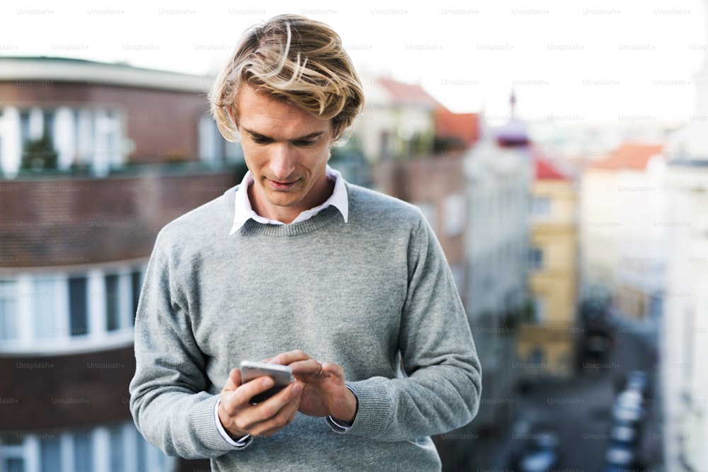 Giovane uomo con smartphone in piedi su una terrazza o balcone in una città, messaggi di testo.