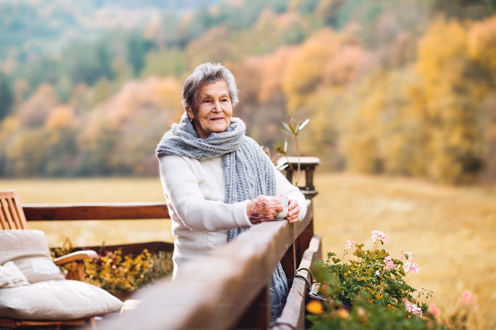An elderly senior woman with a cup of coffee standing outdoors on a terrace on a sunny day in autumn.