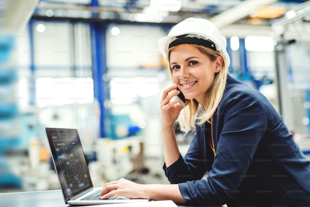 A portrait of a young industrial woman engineer in a factory, using laptop and smartphone.