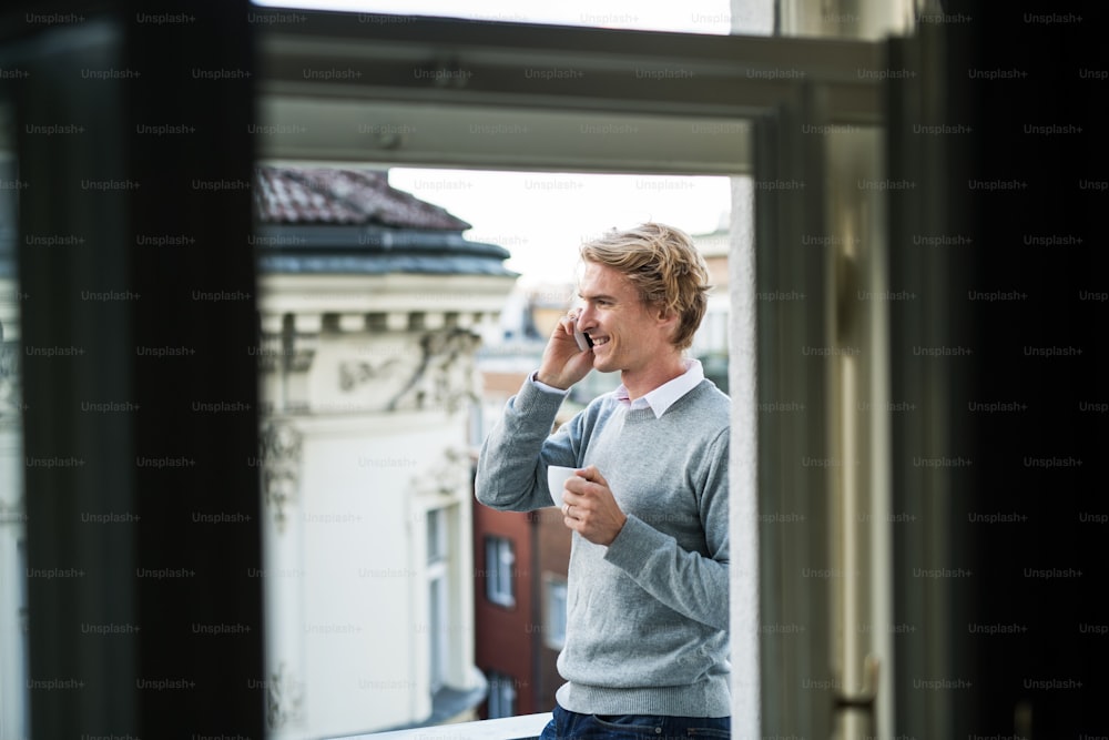 Junger Mann mit Kaffee und Smartphone steht auf einer Terrasse oder einem Balkon in einer Stadt und telefoniert.